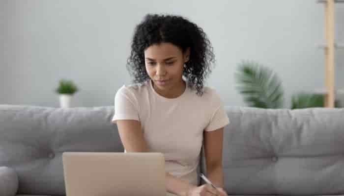 woman sitting on a sofa, looking at a laptop and writing notes