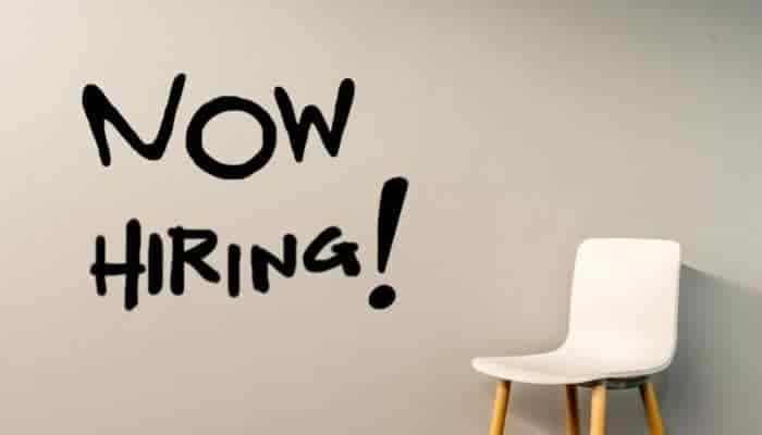 White chair placed against plain cream background. The text Now Hiring is on the wall.