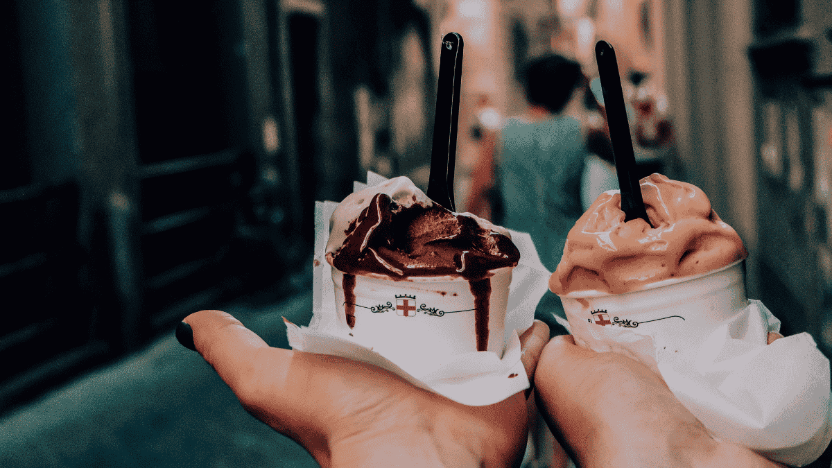 two hands holding small tubs of melting ice cream in a city street